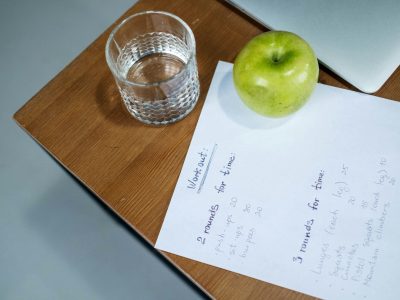 A green apple, glass of water, and workout plan on a wooden desk symbolize healthy living.